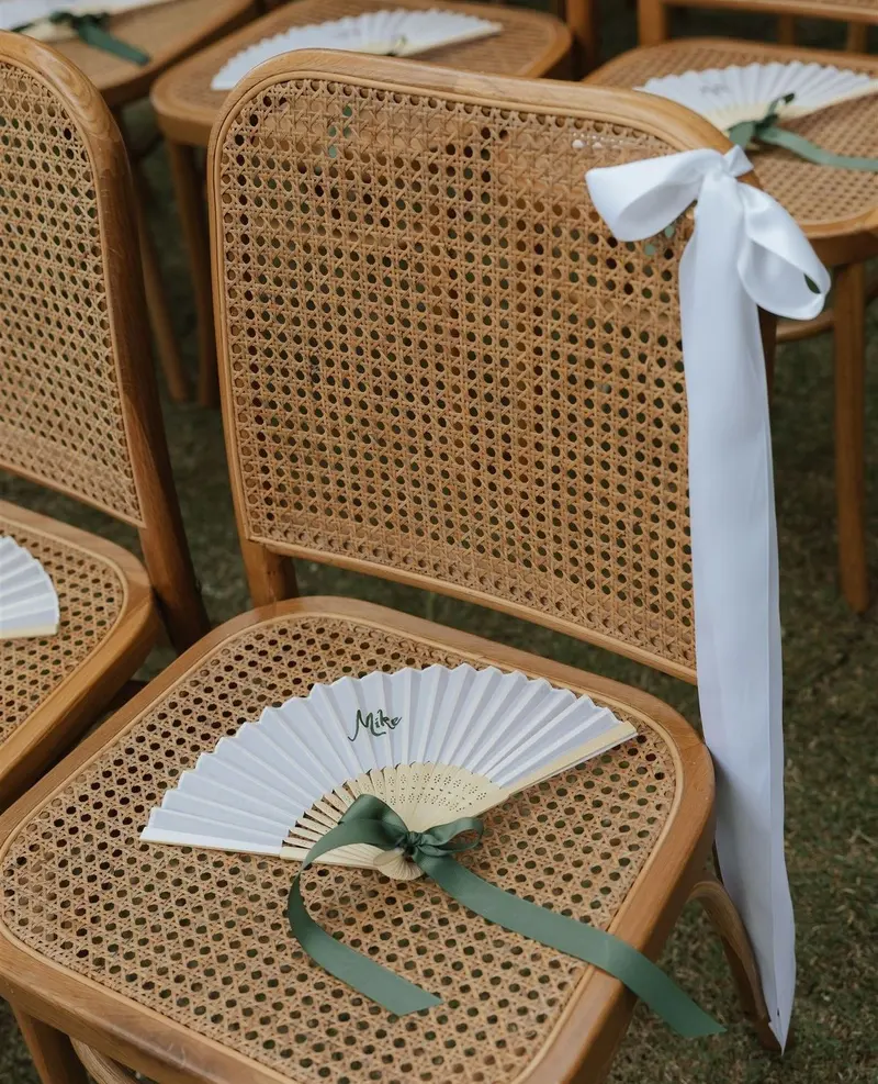 A cane chair at a wedding ceremony with a guest's name on a paper fan tied with a green ribbon