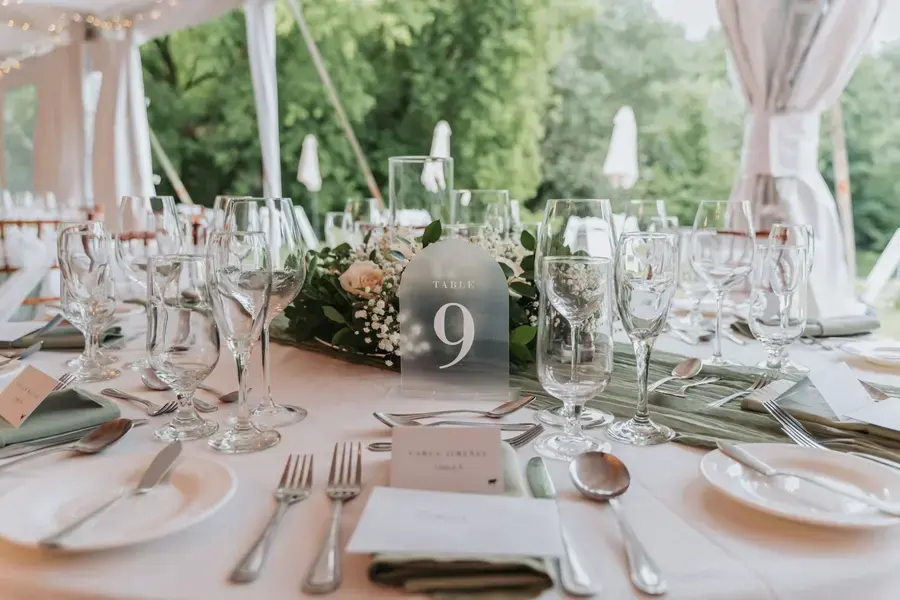 A wedding table with a numbered table sign, place cards, glasses and a flower centrepiece